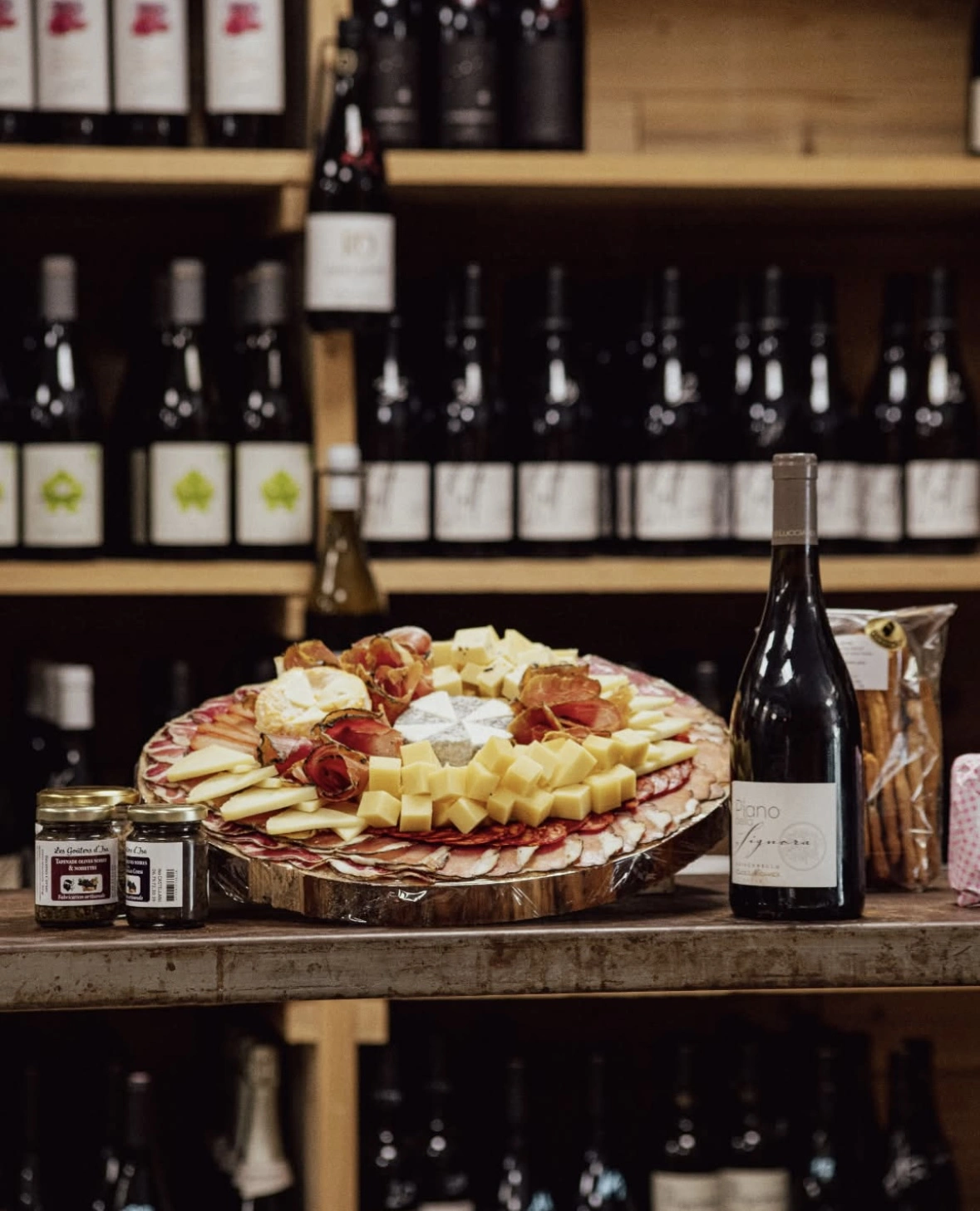 une image d'un plateau de fromage et charcuterie posé sur une table avec une bouteille de vin à coté, et une étagère de vin en fond.